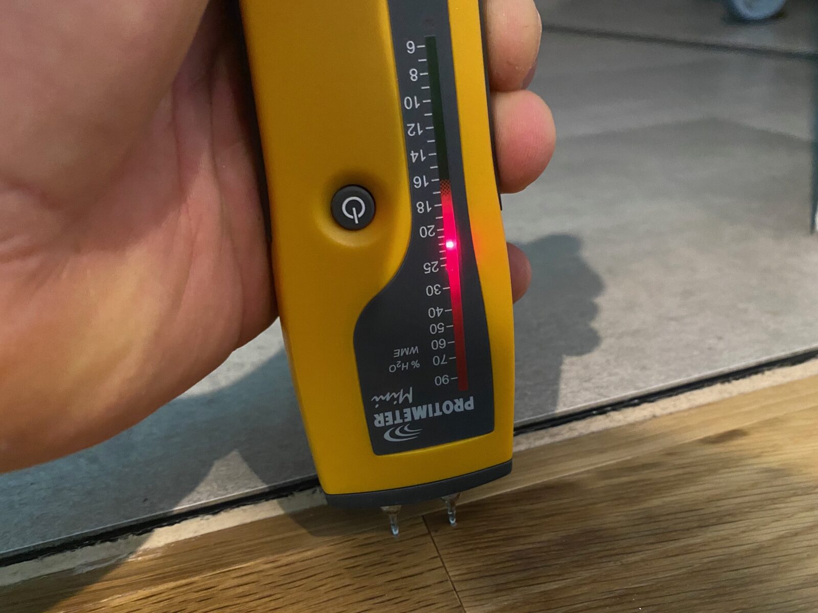 A hand holding a yellow moisture meter with its probes resting at the join between tiled and timber flooring.